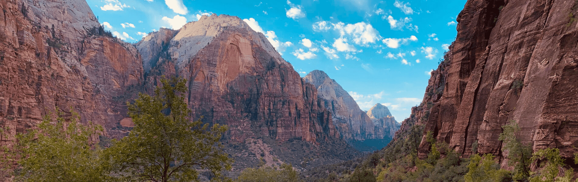 A panoramic view of a wide canyon with towering red and sandstone cliffs, scattered greenery in the valley below, and a bright blue sky filled with scattered white clouds.
