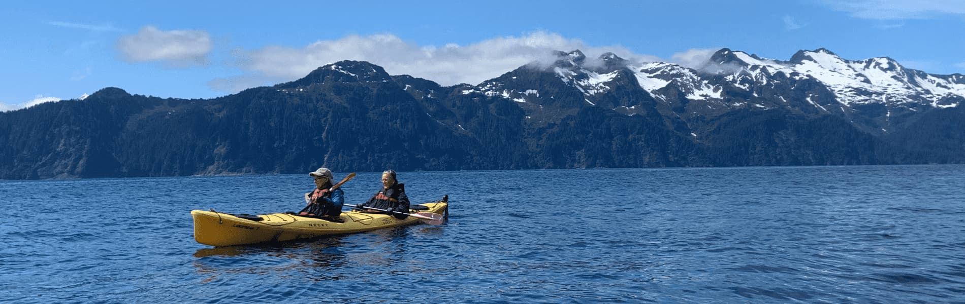 Two people paddling a yellow tandem kayak on a calm blue lake, surrounded by rugged dark mountains with patches of snow under a bright sky.