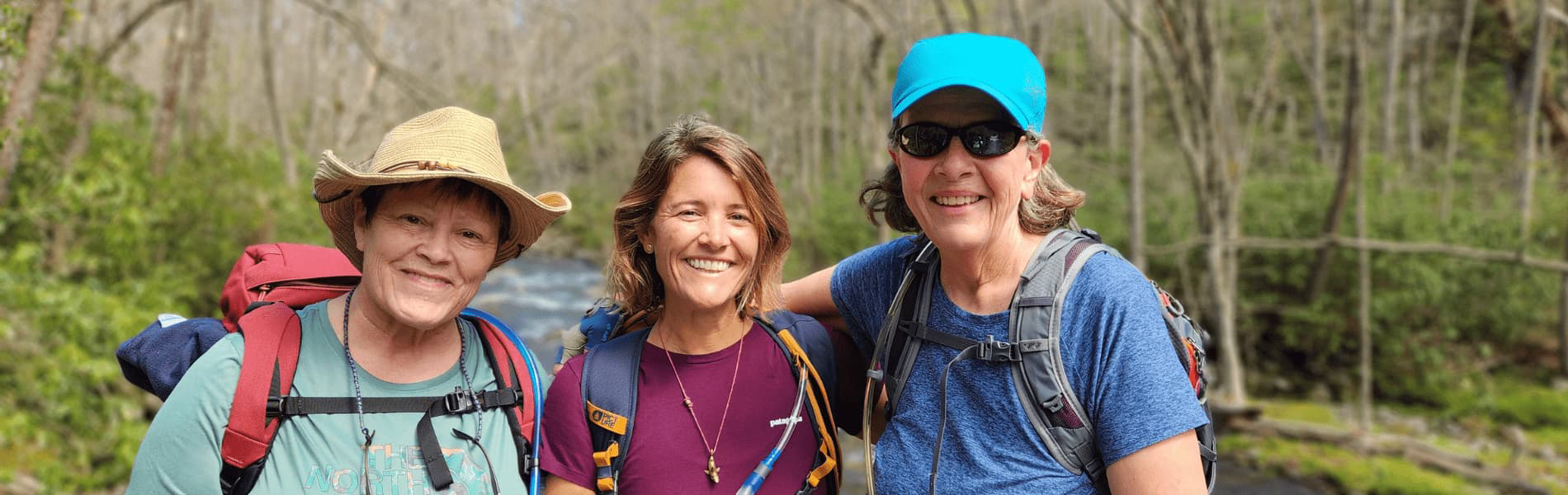 Three hikers standing together on a wooded trail, wearing backpacks and outdoor clothing, with a flowing creek and early‑season forest vegetation in the background.