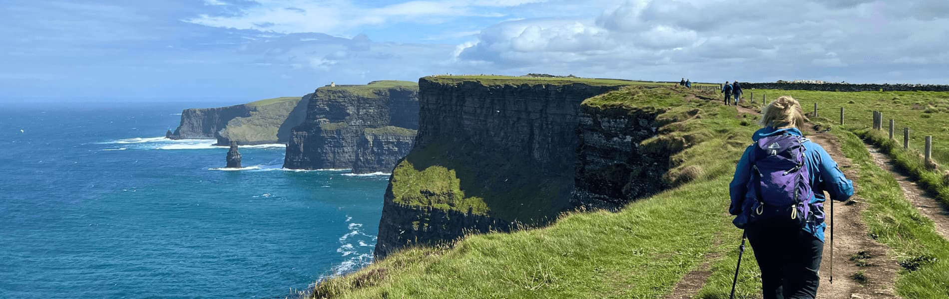 A person hiking along a grassy cliffside trail overlooking tall ocean cliffs and deep blue water under a bright sky with scattered clouds.